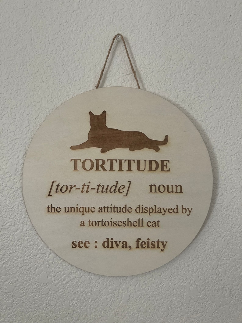 May include: A round wooden sign with a cat silhouette and the word "TORTITUDE" defined as "the unique attitude displayed by a tortoiseshell cat". The sign also says "see: diva, feisty".