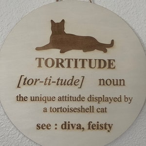 May include: A round wooden sign with a brown silhouette of a cat. The sign reads "TORTITUDE [tor-ti-tude] noun the unique attitude displayed by a tortoiseshell cat see: diva, feisty"