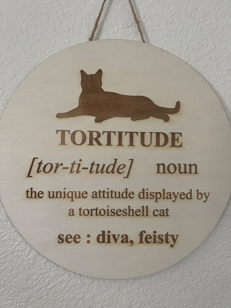 May include: A round wooden sign with a brown silhouette of a cat. The sign reads "TORTITUDE [tor-ti-tude] noun the unique attitude displayed by a tortoiseshell cat see: diva, feisty"