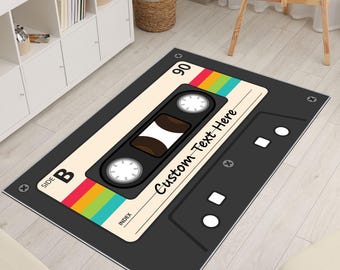 Könnte beinhalten: Ein rechteckiger Teppich, der wie ein Vintage-Kassettenband gestaltet ist. Der Teppich hat einen schwarzen Rand, eine beige Mittelplatte mit dem Text "Custom Text Here" und einen bunten Regenbogenstreifen. Ein dekorativer Artikel.
