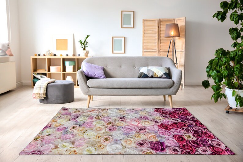 Puede incluir: Una alfombra de &aacute;rea floral con un degradado de rosas rosas, blancas y crema. La alfombra est&aacute; en un ambiente de sala de estar con un sof&aacute; gris y una unidad de estanter&iacute;a de madera.