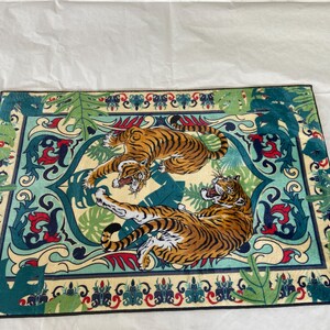 May include: Rectangular rug featuring two tigers in shades of orange and black, set against a cream and teal background with a decorative border. The rug has a jungle theme with stylized leaves and floral elements.