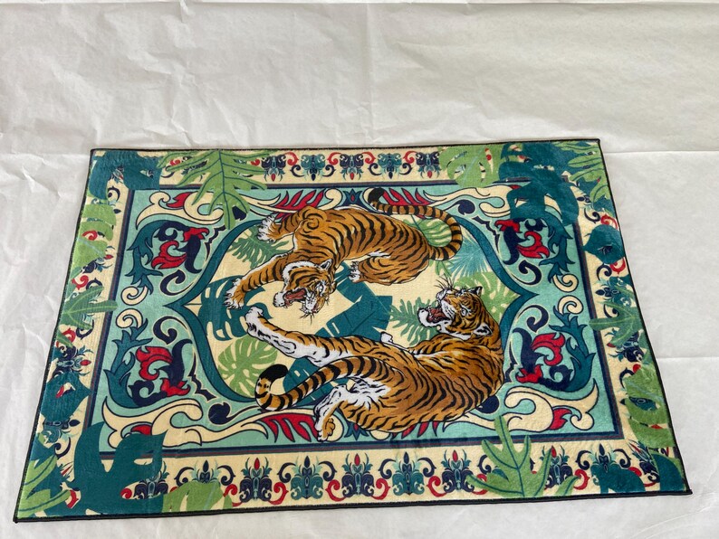 May include: Rectangular rug featuring two tigers in shades of orange and black, set against a cream and teal background with a decorative border. The rug has a jungle theme with stylized leaves and floral elements.