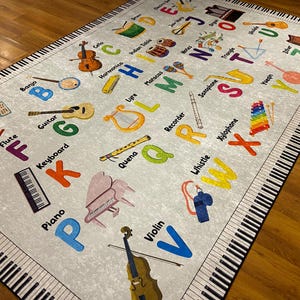 May include: A light gray educational rug featuring the alphabet and musical instruments. The rug has a piano key border and includes illustrations of a piano, violin, guitar, and other instruments. Colorful letters and instrument names are scattered across the rug.