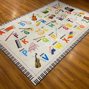 May include: A rectangular rug with a musical theme. The rug features a light gray background with colorful illustrations of musical instruments and the alphabet. The border of the rug is designed to look like piano keys.