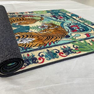 May include: A rolled-up rectangular rug featuring a detailed design of two tigers in a jungle setting. The rug has a cream-colored background with teal, red, and green accents. The tigers are orange and black.