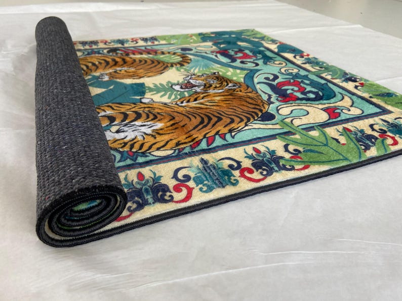 May include: A rolled-up rectangular rug featuring a detailed design of two tigers in a jungle setting. The rug has a cream-colored background with teal, red, and green accents. The tigers are orange and black.