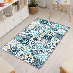 Könnte beinhalten: Rechteckiger Teppich mit einem geometrischen Fliesenmuster in Marineblau, Türkis, Gelb und Weiß. Der Teppich weist verschiedene Blumen- und geometrische Designs innerhalb jeder Fliese auf, wodurch ein Patchwork-Effekt entsteht. Der Teppich ist mit einer dünnen rosa Bordüre eingefasst.