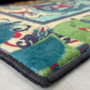 May include: Close-up of a colorful rug with a detailed floral pattern. The rug features a combination of teal, green, red, and cream colors. The edges are finished with a dark gray border, adding a defined shape to the rug.