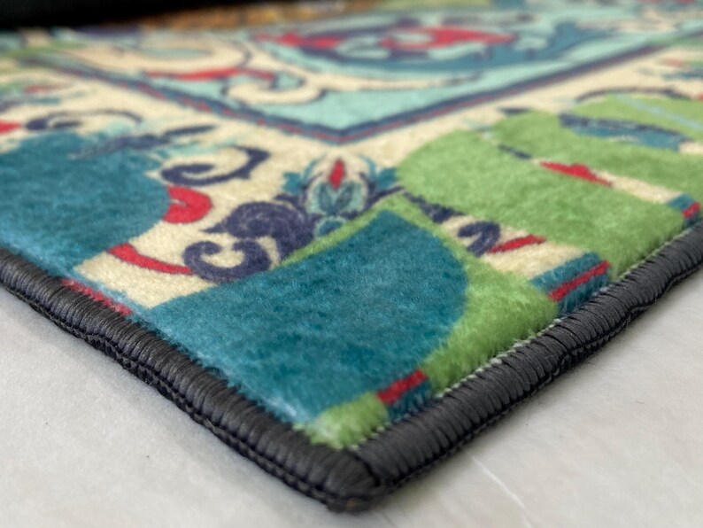 May include: Close-up of a colorful rug with a detailed floral pattern. The rug features a combination of teal, green, red, and cream colors. The edges are finished with a dark gray border, adding a defined shape to the rug.