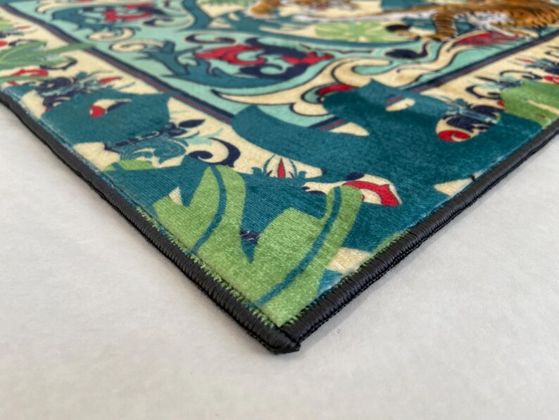May include: Close-up of a decorative rug with a detailed pattern. The rug features a teal, green, and cream color scheme with a black border. The design includes floral and animal motifs.