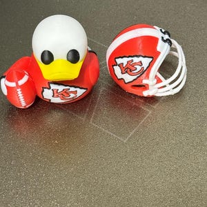 May include: A red and white Kansas City Chiefs football helmet and a red, white, and yellow duck with a football. The duck has the Chiefs logo on its chest and a football in its left wing.