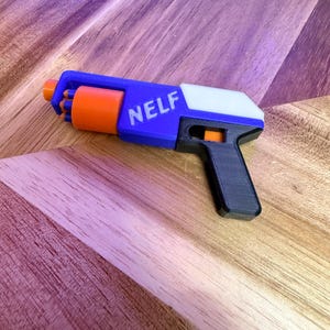 Puede incluir: Una pistola de juguete azul, naranja, blanca y negra con la palabra "NELF" impresa en el lateral. La pistola de juguete está hecha de plástico y tiene un gatillo y un cañón. La pistola de juguete está sobre una superficie de madera.
