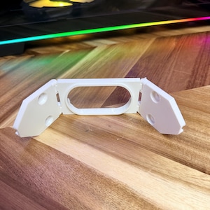 May include: A white, 3D-printed bracket with an oval centre and two angled side pieces. Each side piece has two circular holes. The bracket is on a wooden surface with a computer in the background.