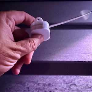 Op de afbeelding: Een wit plastic lichtarmatuurcomponent met een kleine lus aan de bovenkant, tegen een paars oppervlak met textuur. Een dunne witte draad loopt van het armatuur.