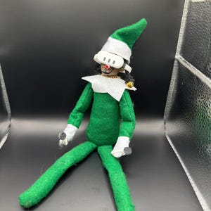 Puede incluir: Una muñeca elfo verde hecha a mano con un gorro puntiagudo, cuello blanco y guantes blancos. El elfo lleva gafas con montura blanca con tres puntos negros y un punto rojo. La muñeca tiene pelo negro y una campana dorada en la oreja.