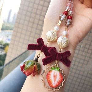May include: A red and white strawberry charm with a gold frame, a red velvet bow, a dried rose, and white and gold beads on a gold chain.
