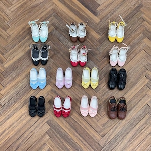 May include: A collection of miniature doll shoes in various styles and colors, including ballet flats, Mary Janes, and ankle-strap heels. The shoes are in shades of blue, pink, yellow, black, red, and brown, with details like bows and lace.