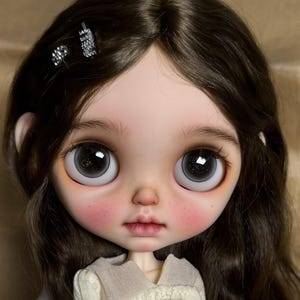 May include: A close-up of a doll's face with large, expressive eyes and dark brown hair. The doll has rosy cheeks and a delicate, neutral-toned lip color. Two small, sparkly hair clips adorn the hair. The doll is wearing a cream-colored sweater.