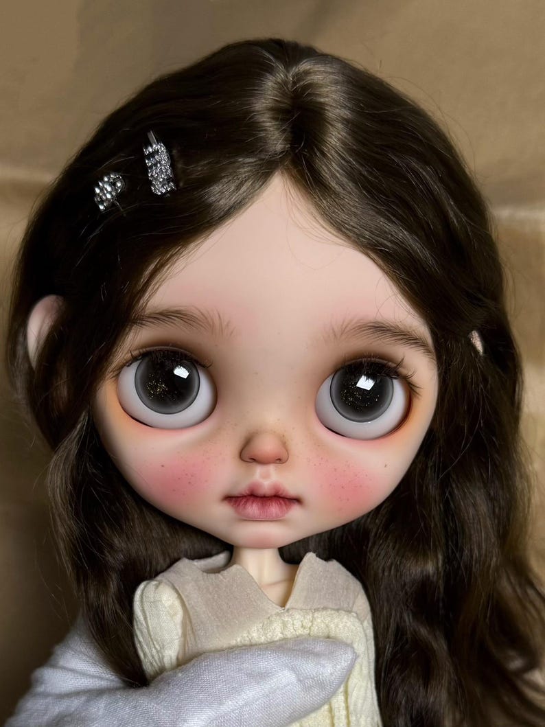 May include: A close-up of a doll's face with large, expressive eyes and dark brown hair. The doll has rosy cheeks and a delicate, neutral-toned lip color. Two small, sparkly hair clips adorn the hair. The doll is wearing a cream-colored sweater.