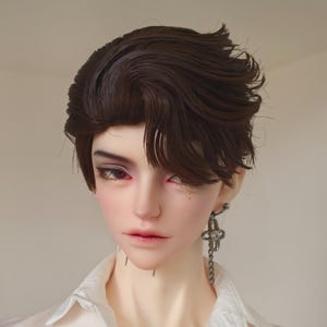 May include: A doll with a realistic face, dark brown hair styled in a side-swept fashion, and a white collared shirt. The doll has detailed makeup with purple eyeshadow and red-toned lips. A silver earring hangs from the left ear.