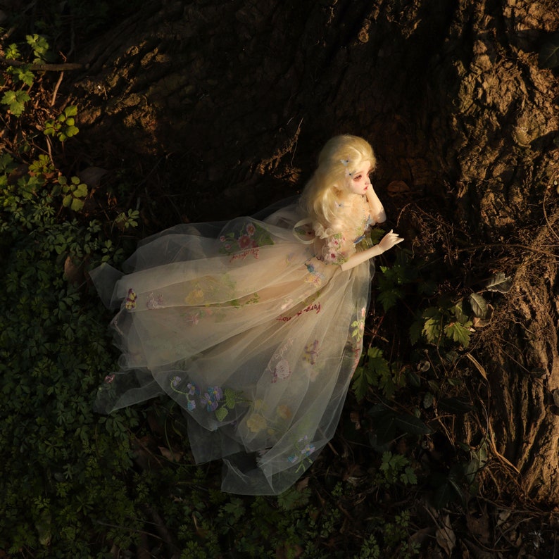 May include: A porcelain doll with blonde hair wearing a white dress with floral embroidery. The doll is lying on a bed of green leaves and brown branches.