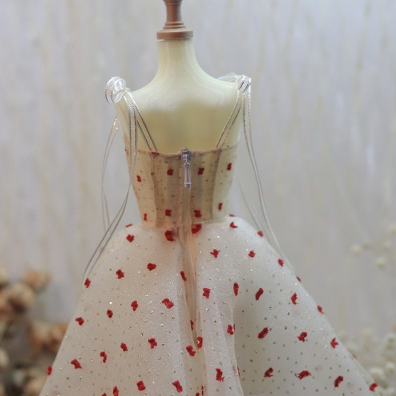 May include: A white tulle dress with red polka dots and a zipper closure in the back. The dress is on a white mannequin.