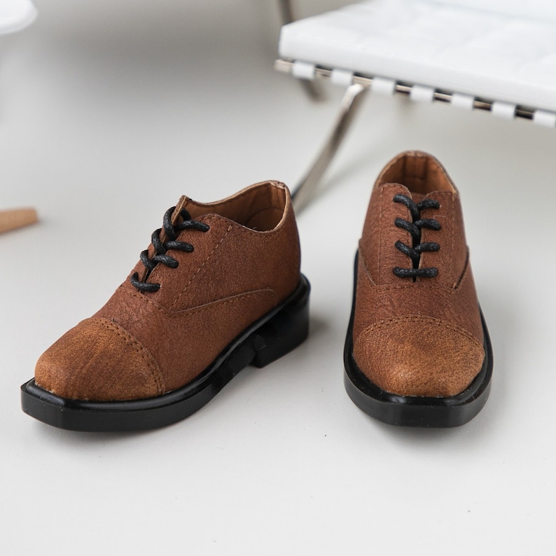 Puede incluir: Un par de zapatos marrones de cuero con cordones negros. Los zapatos tienen una punta redonda y una suela de goma negra.