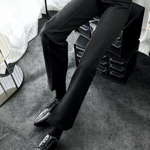 May include: Black trousers with a subtle herringbone weave, worn with black leather dress shoes. The trousers have a slight flare. The shoes are polished and lace up. The outfit is formal and stylish, suitable for a smart occasion.