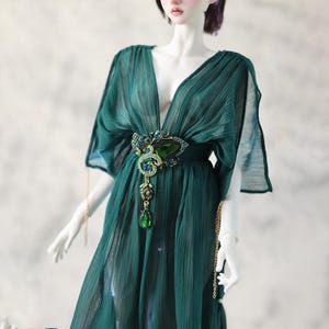 Op de afbeelding: Een pop in een vloeiende, smaragdgroene jurk met een diepe V-hals. De jurk is in de taille getailleerd met een sierlijke broche met een grote groene edelsteen en kleinere blauwe accenten. Gouden kettingen hangen van de broche.