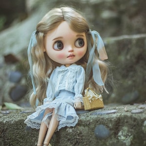 May include: A doll with blonde hair styled in pigtails with blue ribbons. The doll wears a light blue dress with white lace trim and a matching jacket. A small woven basket sits beside the doll.