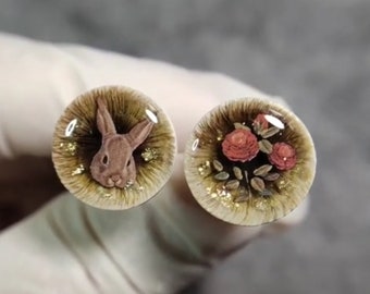 May include: Two round resin cabochons with gold glitter. One features a brown rabbit, the other features three red roses with green leaves.