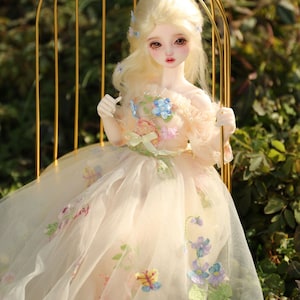 May include: A porcelain doll with blonde hair and blue eyes wearing a white dress with floral embroidery. The doll is sitting on a gold metal chair.