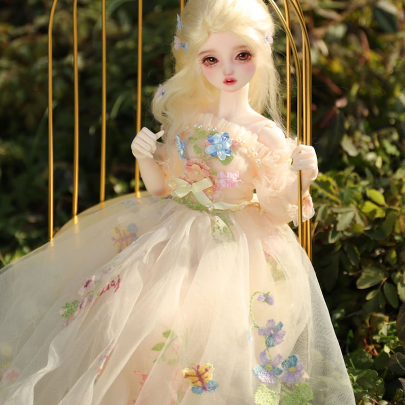 May include: A porcelain doll with blonde hair and blue eyes wearing a white dress with floral embroidery. The doll is sitting on a gold metal chair.