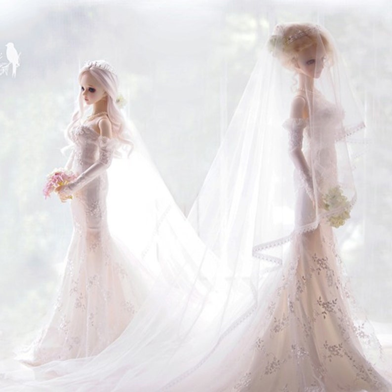 May include: Two white wedding dolls wearing white lace dresses and veils. The dolls are standing side by side with their arms at their sides. The doll on the left is holding a bouquet of pink flowers.