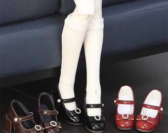May include: Three pairs of doll shoes in varying styles and colors. The shoes include a pair of brown heeled Mary Janes, black Mary Janes, and red Mary Janes. Each pair features a strap and decorative embellishments.