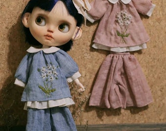 May include: Two doll outfits, one blue and one pink, with embroidered flowers. Both outfits have a collared top and a skirt or trousers.