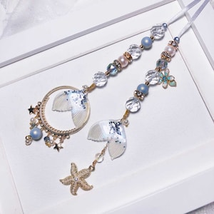 May include: A pair of white and blue mermaid tail charms with gold accents and a starfish charm hanging from a white ribbon with blue and white beads.