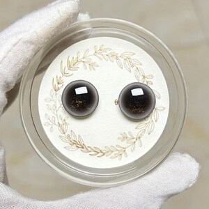 May include: A pair of dark gray stud earrings with a gold flecked interior, presented in a clear plastic case. The earrings sit on a white card with a gold leaf design. The case is held by a gloved hand.