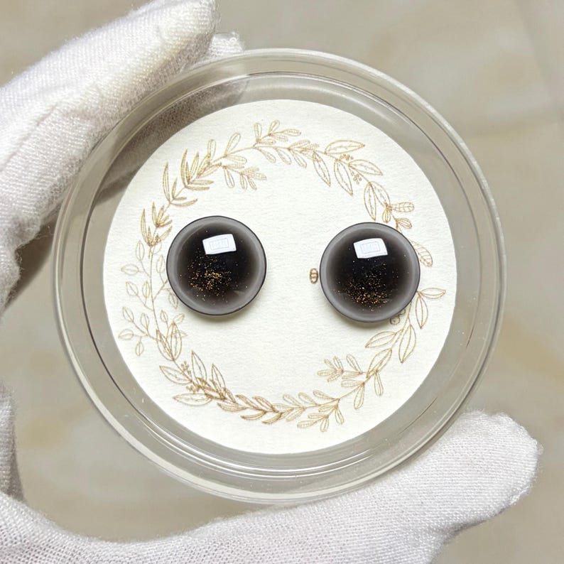 May include: A pair of dark gray stud earrings with a gold flecked interior, presented in a clear plastic case. The earrings sit on a white card with a gold leaf design. The case is held by a gloved hand.