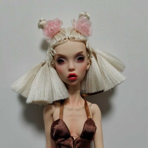 May include: A doll with pale skin, red lips, and dark eye makeup. The doll has white hair styled with two buns adorned with pink flower accents. It wears a brown halter top with gold straps.