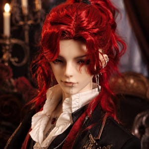 May include: A ball-jointed doll with vibrant red hair styled in a high ponytail. The doll features pale skin, a silver lip chain, and silver ear jewelry. It wears a white ruffled shirt under a black jacket with red trim and a red gemstone pendant.