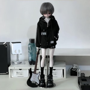 Peut inclure: Une poupée aux cheveux gris vêtue d'une veste noire, d'un short et de bottes. Une guitare électrique noire et une bouteille d'eau sont à côté de la poupée. La poupée est sur une surface blanche.