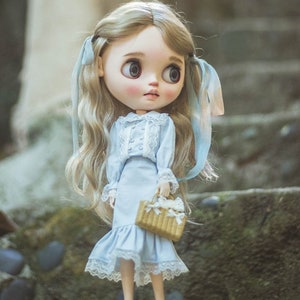 May include: A doll with long, wavy blonde hair and blue ribbons. The doll wears a light blue dress with lace trim and a matching skirt. She holds a small woven basket with white accents. The doll has large eyes and rosy cheeks.