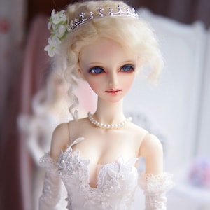 May include: A porcelain doll with blonde hair styled in a bun, wearing a white lace dress and a silver crown. The doll has blue eyes and a pearl necklace.