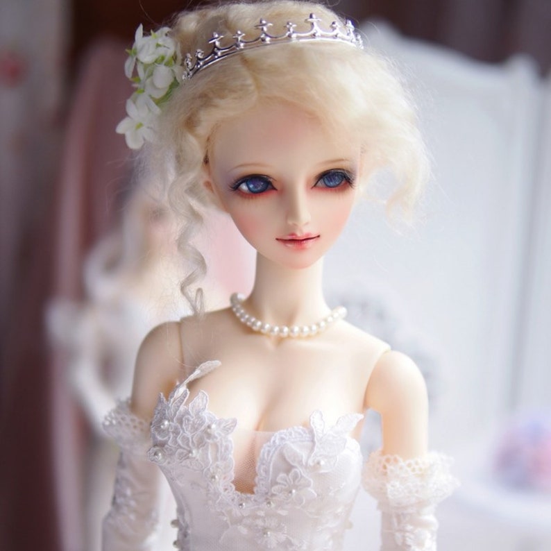 May include: A porcelain doll with blonde hair styled in a bun, wearing a white lace dress and a silver crown. The doll has blue eyes and a pearl necklace.