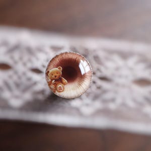 May include: A round, brown and white ring with a small teddy bear image. The ring has a silver-colored border and is set against a white lace background. The teddy bear is light brown with a white belly.