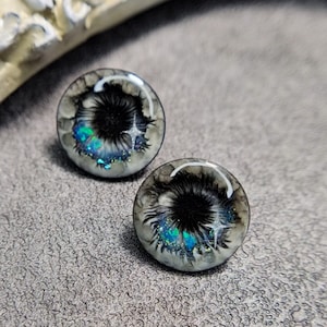 Op de afbeelding: Een paar ronde, hars oorbellen met een zwart-wit swirl-ontwerp en iriserende glitteraccenten. De oorbellen hebben een witte achtergrond met een zwarte swirl in het midden en iriserende glitter die eroverheen is gestrooid.