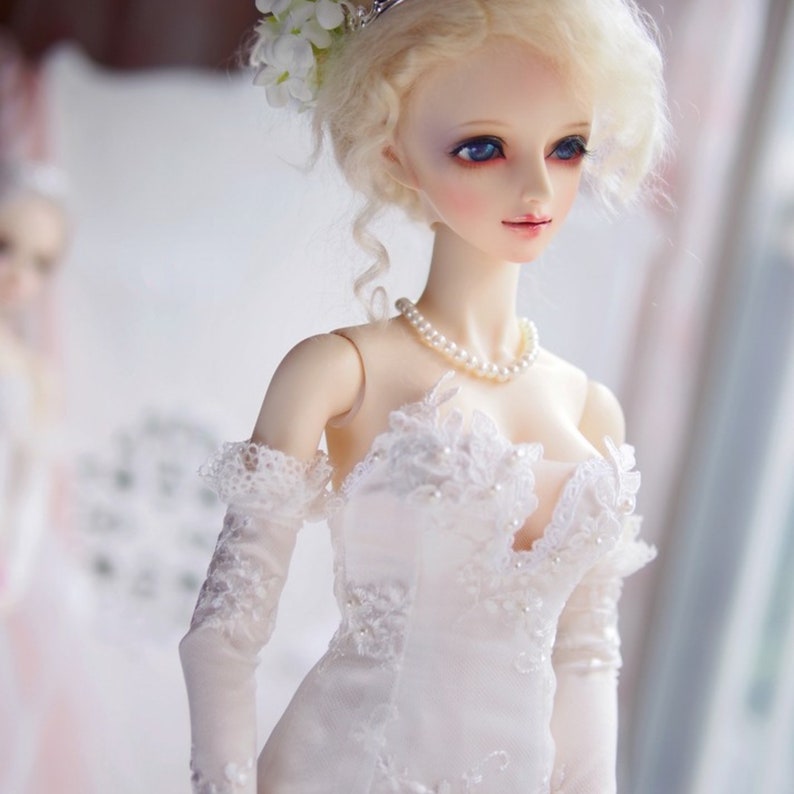 May include: A porcelain doll wearing a white lace wedding dress with a pearl necklace. The doll has blonde hair styled in a bun with white flowers and blue eyes.