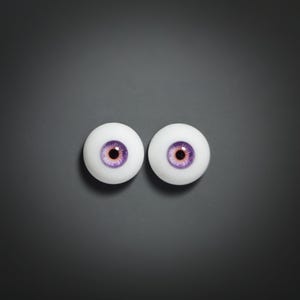Könnte beinhalten: Zwei weiße Puppenaugen mit violetten Iris und dunkelbraunen Pupillen, vor einem dunkelgrauen Hintergrund. Die Augen sind rund und scheinen aus einem glatten, glänzenden Material zu bestehen. Die Gesamtästhetik ist sauber und einfach.